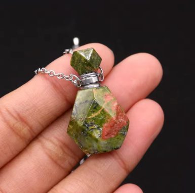 A close-up of a hand holding a small pendant necklace made from a faceted green stone with orange-red inclusions and a silver chain.