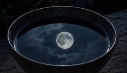A dark ceramic bowl filled with water, reflecting a full moon and cloudy night sky onto a dark wooden surface.