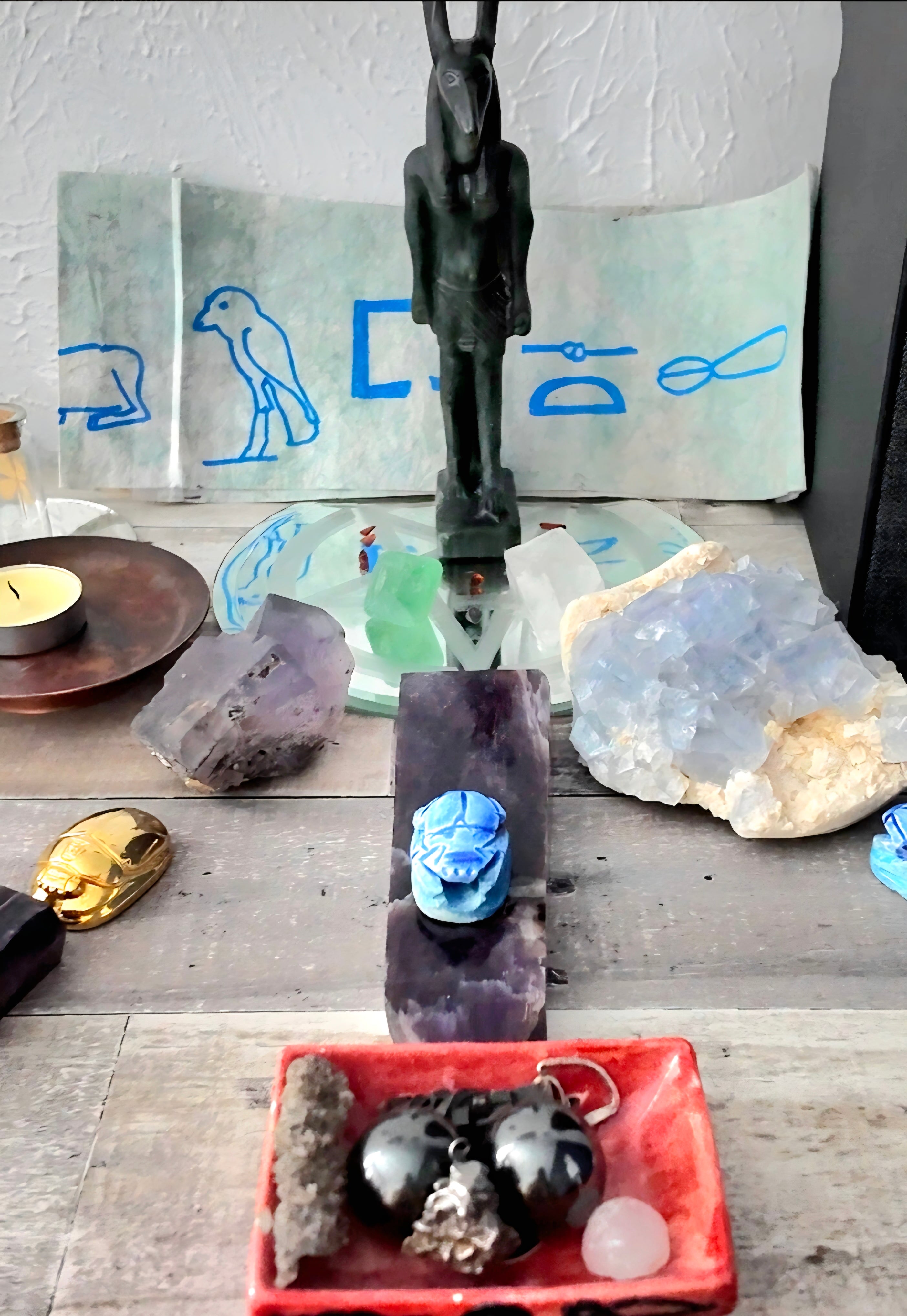 A tabletop display featuring a black metal Egyptian pharaoh figurine, various raw crystals, a blue carved skull, and a red tray holding dark polished stones, set against a light-colored wall with blue line drawings.