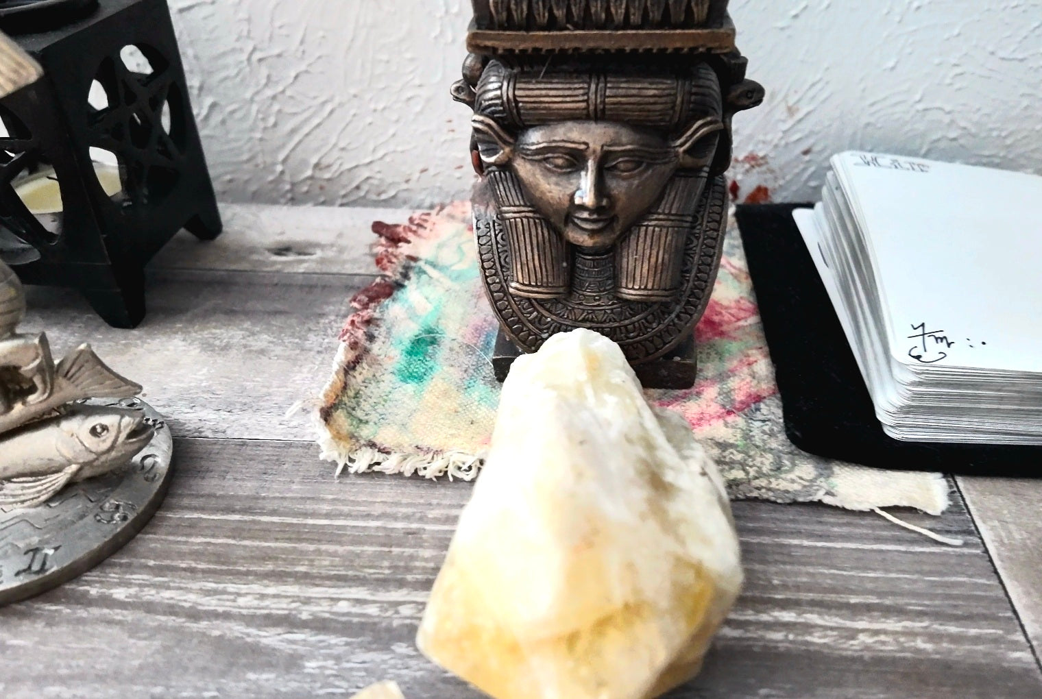 A decorative bronze-toned metal bust sculpture of a face with an ornate headdress, displayed on a rustic wooden surface alongside a large pale crystal, a folded multicolor cloth, a stack of white cards, and a black lantern.
