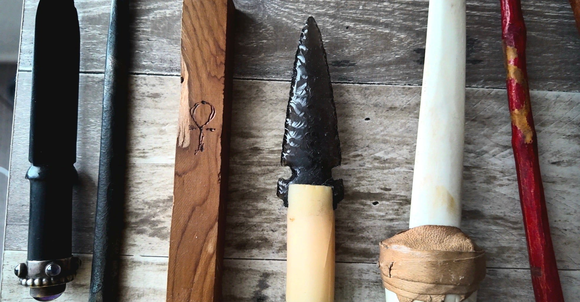 A collection of handcrafted knives and tools laid out on a rustic wooden surface, including a fixed-blade knife with a black textured blade and cream handle, a wooden scabbard with a carved symbol, and other blades.