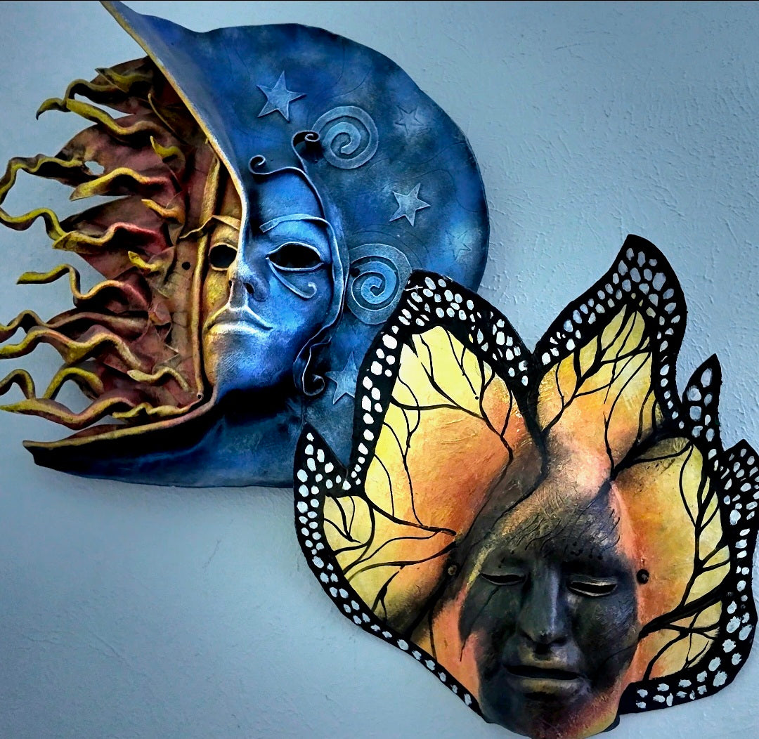 Two decorative wall art masks displayed against a light blue wall. One mask is blue with a moon and star motif, and the other is yellow and orange with butterfly wings and a face design.