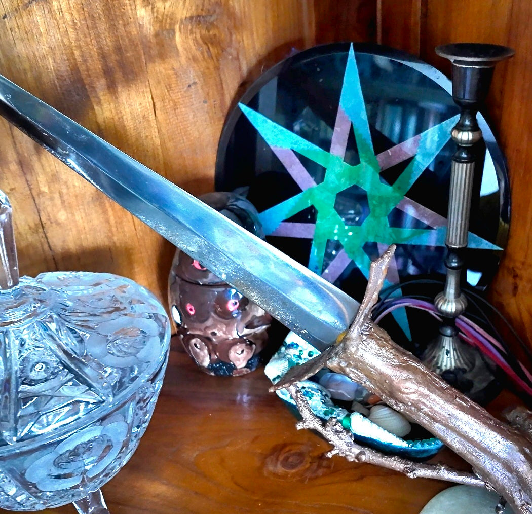 A decorative fantasy sword with a silver blade and a textured brown hilt, displayed on a wooden shelf alongside a glass bowl, a star-shaped mirror, and other mystical objects.
