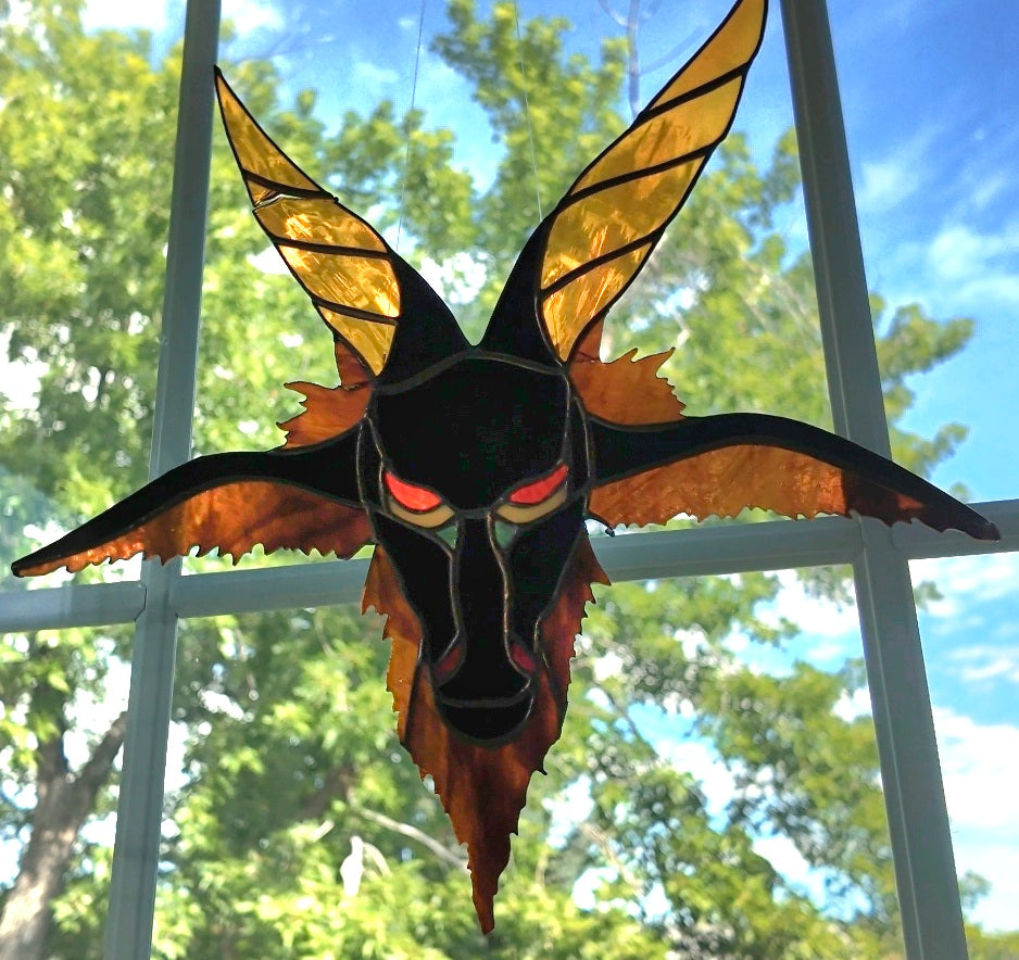 A stained glass suncatcher shaped like a horned demon or beetle, featuring gold and orange wings, a black face, and glowing red eyes, suspended in front of a window with green trees and blue sky visible.