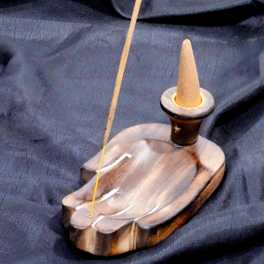 A wooden incense holder shaped like a hand, with a cone-shaped incense stick inserted into a metal stand, set against a dark draped fabric background.