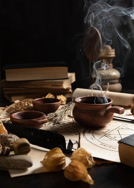 Image of incense burning on a table with esoteric papers and herbs strewn about and a lamp and books stacked.