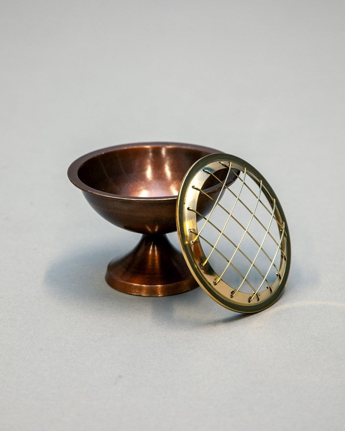A small copper-colored pedestal bowl with a round gold-toned metal grate lid leaning against it, photographed on a plain light gray studio background.
