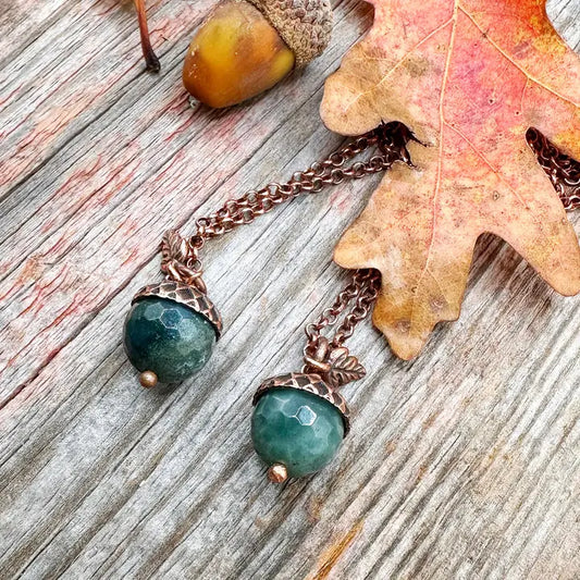 Pair of earrings with green gemstones and copper accents on a wooden surface with an acorn and leaf.