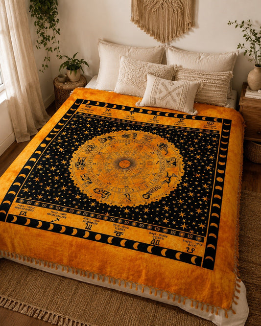 A bed dressed in a large zodiac-themed blanket featuring a golden astrological wheel, moon phases, and stars on a black background, styled in a warm, boho bedroom with cream pillows and plants.