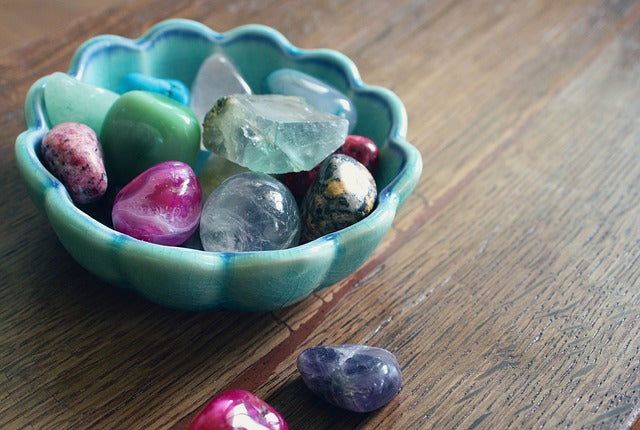 Blue ceramic bowl with sculpted flower like top and many different types of crystals and stones in it and next to the bowl on a wooden table.