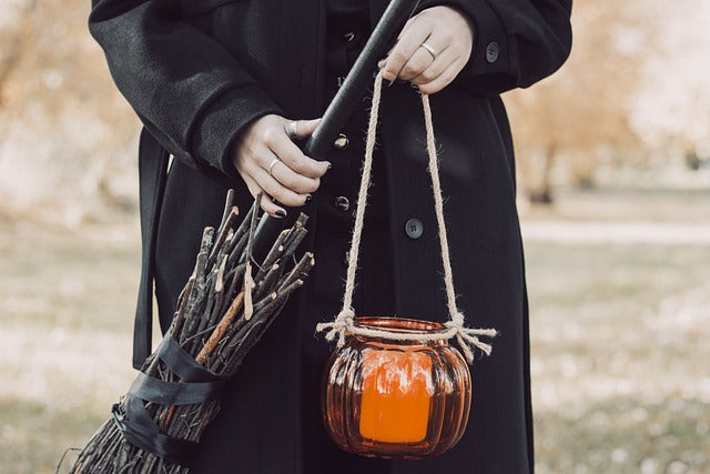 Witch in black holding an orange glass lantern with a candle in it and a pomade broom they have crafted.